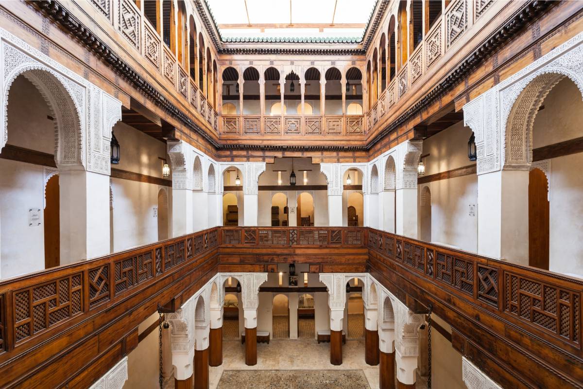 Elegant Moroccan riad courtyard featuring symmetrical Islamic arches, ornate wooden mashrabiya screens, carved balconies, and a central reflecting pool with natural skylight illumination